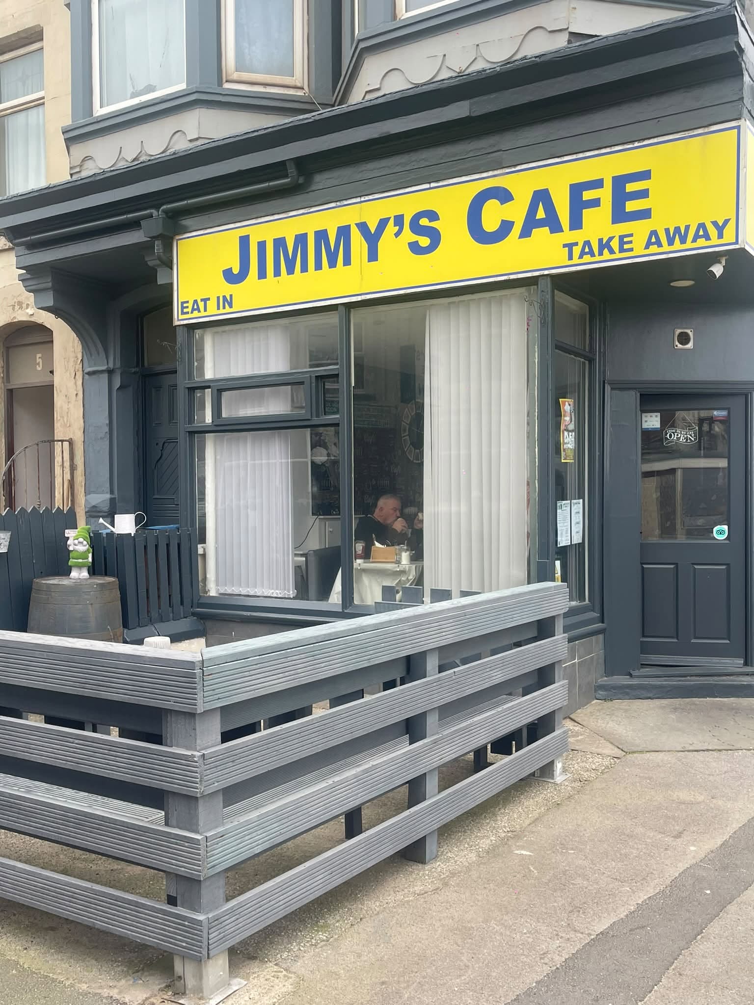 Jimmy’s Cafe’s yellow sign with blue lettering, grey frontage, complete entrance, wooden enclosure and pavement